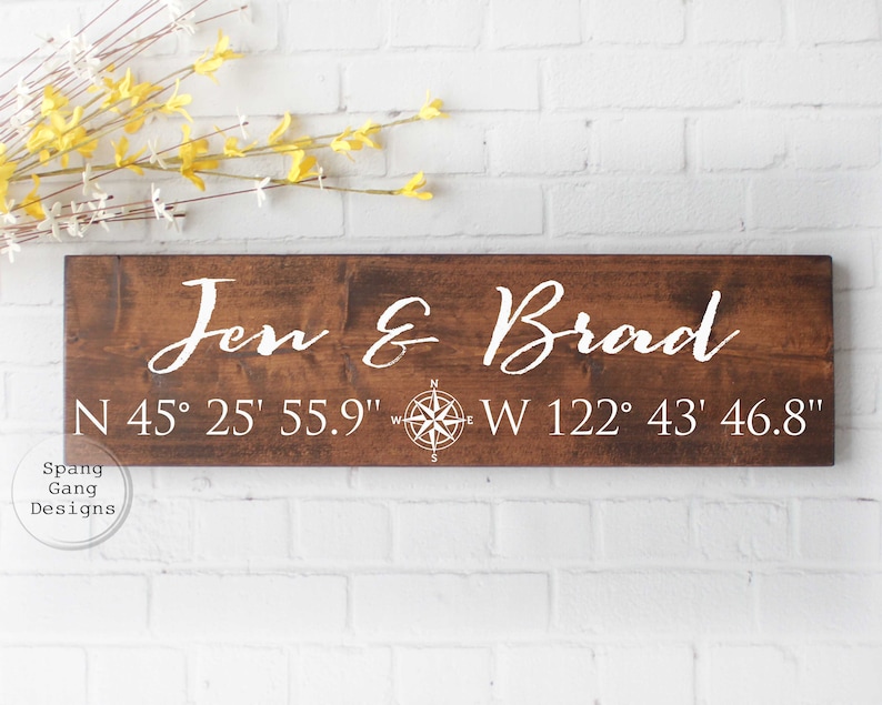 May include: A wooden sign with the names "Jen & Brad" and coordinates "N 45&deg; 25' 55.9" W 122&deg; 43' 46.8" written in white paint. A compass rose is also included on the sign.