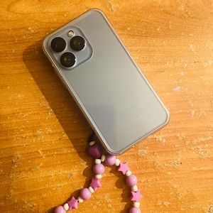 Peut inclure: Un smartphone bleu clair dans une coque de protection transparente, doté d'un système de caméra à trois objectifs. Une dragonne de téléphone perlée est attachée, composée de perles sphériques blanches, de perles sphériques violettes et d'étoiles violettes.