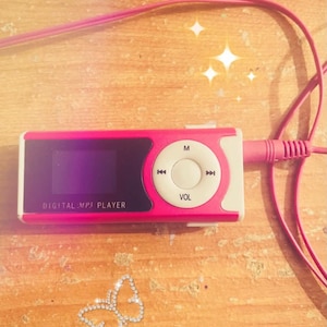 May include: A rectangular digital MP3 player with a hot pink and white casing, a black screen, and a circular white control pad featuring M, VOL, and track navigation buttons. A pink headphone cable is plugged into the side of the device.