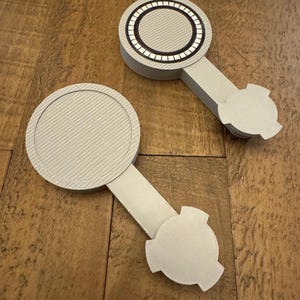 May include: Two light gray, 3D-printed plastic objects, each featuring a circular head with a textured, diagonal-lined surface attached to a rectangular stem and a three-pronged base. One circular head includes a black ring with white segments.