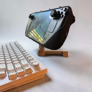 May include: A black handheld gaming console with a screen displaying a game, mounted on a beige adjustable stand, positioned next to a white mechanical keyboard featuring a prominent circular volume knob on the top right corner of the frame.