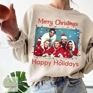 May include: A beige crewneck sweatshirt featuring a rectangular graphic of four individuals wearing red and white holiday attire. The text Merry Christmas and Happy Holidays is printed in red above and below the image, framed by string lights.