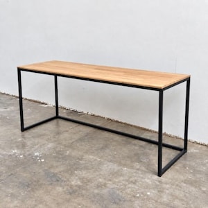 May include: A rectangular table featuring a light brown wooden top with a visible grain pattern. The table is supported by a black metal frame constructed from thin, square-profile tubing that forms a continuous open base structure.