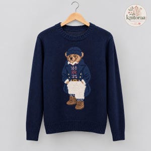 Può includere: Un maglione blu navy a maniche lunghe con girocollo caratterizzato da un motivo centrale in maglia intarsia raffigurante un orso marrone che indossa un berretto blu, un blazer blu, un gilet a motivi, pantaloni bianchi e stivali marroni.