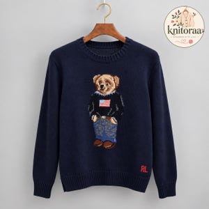 Peut inclure: Un pull en tricot bleu marine à manches longues avec un motif central représentant un ours en peluche portant un pull noir avec un drapeau américain et un jean bleu. Le pull présente un col rond, des poignets et un ourlet côtelés, avec RL en rouge.