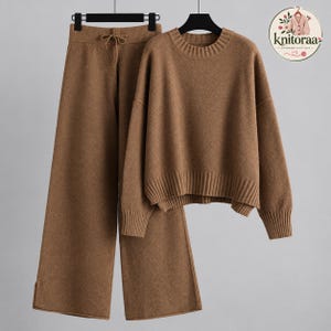 Op de afbeelding: Een bruine tweedelige gebreide set bestaande uit een trui met lange mouwen met een geribbelde ronde hals en boorden, en een wijde broek met een elastische tailleband met trekkoord. De set bevat een rond logo met de tekst knitoraa.