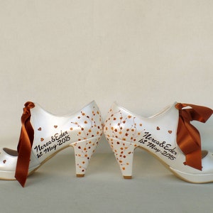 May include: A pair of white high-heeled shoes with brown ribbon bows. The shoes are decorated with brown painted leaves and hearts. The text "Nerea & Eder 1st May 2015" is written on the shoes.