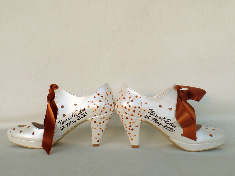 May include: A pair of white high-heeled shoes with brown ribbon bows. The shoes are decorated with brown painted leaves and hearts. The text "Nerea & Eder 1st May 2015" is written on the shoes.