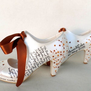 May include: A pair of white satin wedding shoes with a brown ribbon. The shoes are decorated with a floral design and the text "The Book of Love is Long and Boring and Written Very Long Ago. It's Full of Flowers and Heart Shaped Breasts and Things We're All Too Young To Know I Love You" and "Nerea & Eder 1st May 2015".