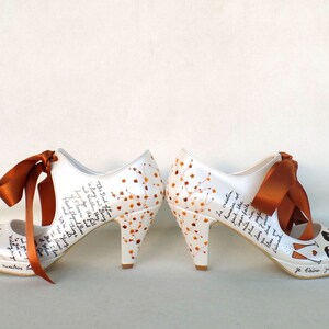 May include: A pair of white high-heeled shoes with orange ribbons. The shoes are decorated with a floral design and handwritten text. The text includes the words "The Book of Love" and "Je t'aime jusqu'&agrave; la lune et retour".