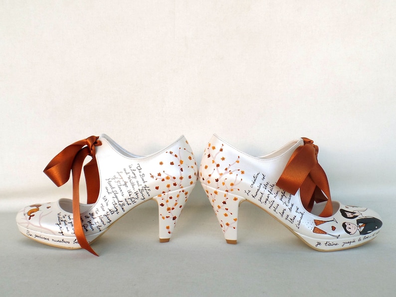 May include: A pair of white high-heeled shoes with orange ribbons. The shoes are decorated with a floral design and handwritten text. The text includes the words "The Book of Love" and "Je t'aime jusqu'&agrave; la lune et retour".