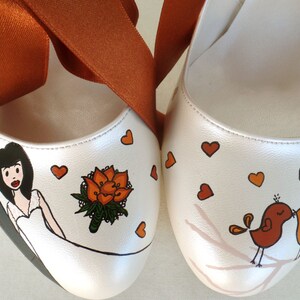 May include: A pair of white wedding shoes with orange ribbon ties. One shoe is painted with a bride and groom, a bouquet of orange flowers, and hearts. The other shoe is painted with two birds kissing on a branch with hearts.