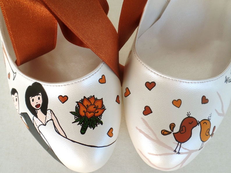 May include: A pair of white wedding shoes with orange ribbon ties. One shoe is painted with a bride and groom, a bouquet of orange flowers, and hearts. The other shoe is painted with two birds kissing on a branch with hearts.
