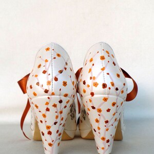 May include: A pair of white satin high-heeled shoes with orange floral designs and orange satin ribbon ties.