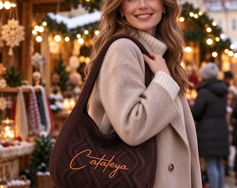 Peut inclure: Un sac à bandoulière en crochet marron foncé avec un motif en tricot torsadé texturé et le nom Cataleya brodé en lettres cursives orange sur le devant. Le sac possède une large bandoulière et est porté sur un manteau en laine beige.