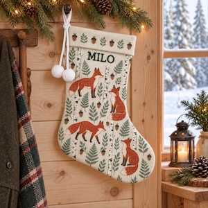 Könnte beinhalten: Ein cremefarbener Weihnachtsstrumpf mit einem wiederkehrenden Muster aus orangefarbenen Füchsen, grünen Farnen und braunen Eicheln. Das Bündchen ist mit dem Namen MILO in grüner Schrift personalisiert und mit einer Reihe von Eicheln verziert.