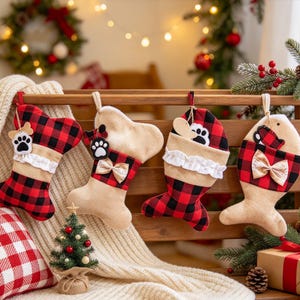 May include: Four Christmas stockings hanging on a wooden rack. Two are shaped like bones and two like fish, featuring a red and black buffalo plaid pattern, beige fabric sections, white ruffled trim, and small decorative bows and paw print tags.