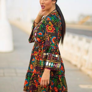 Puede incluir: Un vestido corto negro con un vibrante estampado floral y geométrico multicolor. El vestido tiene mangas largas acampanadas adornadas con adornos de espejo rectangulares y detalles metálicos dorados en los puños y el escote.