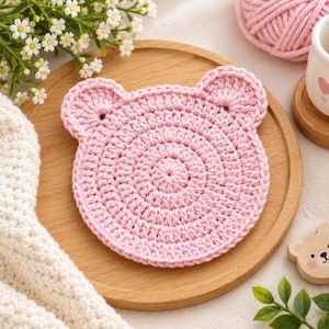 May include: A circular, light pink crochet coaster featuring two small, rounded ears at the top, creating a bear-like silhouette. The coaster is crafted with a dense, concentric stitch pattern, resulting in a textured, flat surface.
