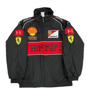 May include: A black racing jacket featuring a prominent red horizontal stripe across the chest with the word Ferrari embroidered in black. The jacket includes various sponsor logos such as Shell, OMP, Bridgestone, and F-1, along with the Scuderia Ferrari emblem and the prancing horse logo on the sleeves.