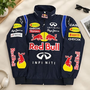 Pode incluir: Um casaco de corrida azul-marinho escuro com fecho de correr, apresentando vários logótipos de patrocinadores, incluindo Red Bull, Infiniti, Pepe Jeans, Geox e Pirelli. O casaco tem gola alta, mangas compridas com punhos elásticos e gráficos de touros vermelhos.