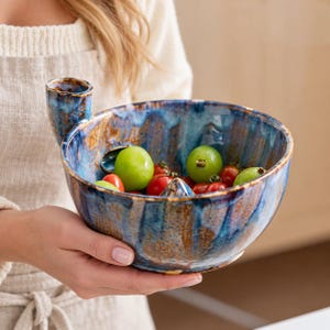 May include: A round ceramic bowl with a blue and brown glazed finish, featuring a small integrated handle on the rim and a central cone-shaped protrusion. The bowl contains a mix of fresh green grapes and small red cherry tomatoes.