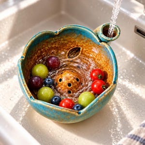 May include: A ceramic berry colander bowl with a teal blue exterior and a speckled brown interior. The bowl features a central drainage dome and a side handle, currently filled with fresh red tomatoes, green grapes, and blueberries.