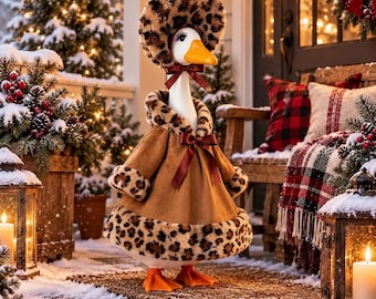 May include: A white goose figurine wearing a brown coat with leopard print trim on the collar, cuffs, and hem, and a matching leopard print bonnet. The outfit features a dark red ribbon bow at the neck and a matching bow on the front of the coat.