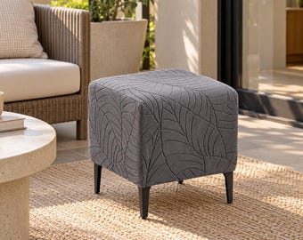 Puede incluir: Un taburete cuadrado con tapicería de tela gris oscuro y un patrón de hojas texturizado en relieve. La pieza se apoya sobre cuatro patas negras delgadas y rectas, proporcionando una base estable para este mueble funcional.