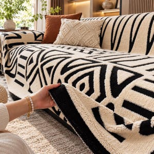 May include: A cream-colored sofa cover featuring a bold, raised black geometric pattern. The fabric has a textured, tufted appearance with a thick, soft pile. The cover drapes over the sofa, showcasing the high-contrast design and tactile surface.