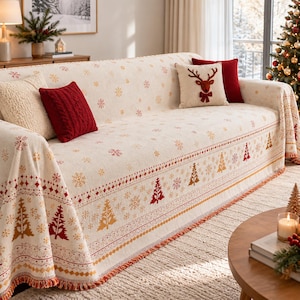 May include: A cream-colored sofa throw blanket featuring a repeating pattern of red and gold Christmas trees and snowflakes. The blanket has a fringed edge and is decorated with two solid red throw pillows and one cream pillow with a reindeer.