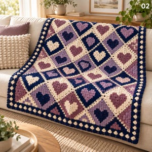 May include: A crochet blanket featuring a repeating diamond pattern of hearts in shades of navy blue, cream, and various tones of purple. The blanket has a thick, dark navy blue border with a textured, scalloped edge along the entire perimeter.