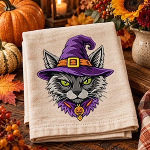 May include: A beige kitchen towel featuring an embroidered design of a gray cat wearing a purple witch hat with an orange band and a gold buckle. The cat has bright green eyes and wears a purple collar with a small orange jack-o'-lantern charm.
