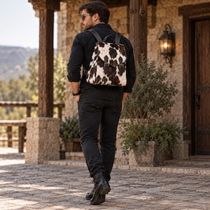 Puede incluir: Una mochila con estampado de piel de vaca con manchas blancas y negras, una solapa superior con dos correas de cuero marrón oscuro y hebillas metálicas. La bolsa tiene correas de hombro de cuero marrón oscuro y un asa superior.