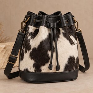 May include: A bucket bag featuring a black and white cow print hide pattern on the main body, accented with smooth black leather trim at the base, top, and drawstring closure. It includes an adjustable black leather shoulder strap with brass hardware.