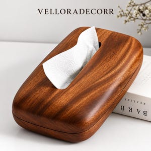 Puede incluir: Una caja de madera rectangular para pañuelos con un acabado de grano marrón oscuro y bordes redondeados. Un pañuelo blanco con un sutil patrón en relieve sobresale de la abertura ovalada central. El texto VELLORADECORR es visible arriba.