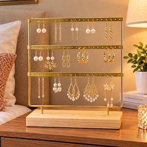 May include: A gold-toned metal jewelry organizer stand with three horizontal bars featuring evenly spaced holes for hanging earrings. The metal frame is supported by a rectangular light-colored wooden base, displaying various dangling earrings.
