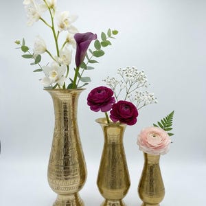 Puede incluir: Tres jarrones de latón de tamaños graduados con patrones florales grabados. El más alto contiene orquídeas blancas y eucalipto. El mediano contiene dos ranúnculos magenta y paniculata. El más pequeño contiene un ranúnculo rosa pálido y helecho.