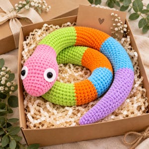 Könnte beinhalten: Ein geringeltes Häkel-Schlangenspielzeug mit segmentiertem Design in abwechselnden Farben: Pink, Limettengrün, Orange, Blau und Lavendel. Die Schlange hat zwei große, runde Sicherheitsaugen aus weißem und schwarzem Kunststoff am pinken Kopf.