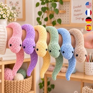 Könnte beinhalten: Eine Sammlung von sechs gehäkelten Schlangenspielzeugen in Rosa, Lila, Gelb, Hellgrün, Blau und Beige. Jede Schlange hat eine strukturierte Garnoberfläche, einen abgerundeten Kopf und zwei kleine schwarze Perlenaugen, die zu einer Schlaufe geformt sind.