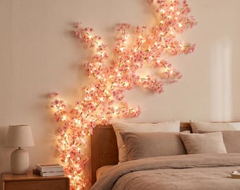 Puede incluir: Una rama decorativa montada en la pared con numerosas flores de cerezo de color rosa claro, iluminadas por pequeñas luces LED de color blanco cálido. La rama se extiende vertical y horizontalmente, creando un despliegue floral brillante.