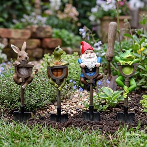 May include: Four garden stakes featuring sculpted figures of a brown rabbit, a green turtle, a gnome with a red hat and white beard, and a green frog. Each figure is attached to a miniature shovel handle, with the shovel blade at the base.