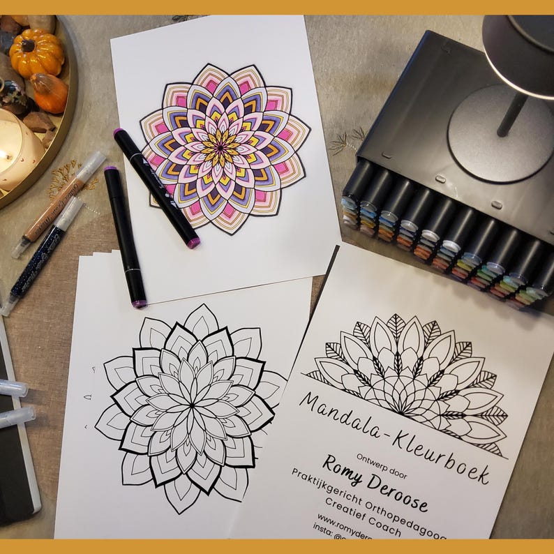 Op de afbeelding: Een kleurboek getiteld Mandala-Kleurboek door Romy Deroose, met twee mandala-illustraties, &eacute;&eacute;n ingekleurd in roze, geel en paars, en &eacute;&eacute;n zwart-wit omtrek. Een zwarte stift en een set stiften in een donkere etui zijn inbegrepen.