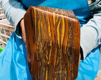 May include: A large, polished slab of tiger's eye gemstone featuring a prominent chatoyant pattern of golden-yellow and deep brown vertical bands. The stone has a smooth, reflective surface with a rectangular shape and rounded edges.