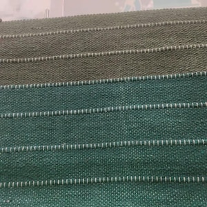 Op de afbeelding: Een stoffenstaalboek met horizontale stroken van getextureerd materiaal in tinten olijfgroen en diep teal. Elke strook wordt gescheiden door een dunne, witte, horizontale stiklijn, wat zorgt voor een gelaagd, rasterachtig uiterlijk.