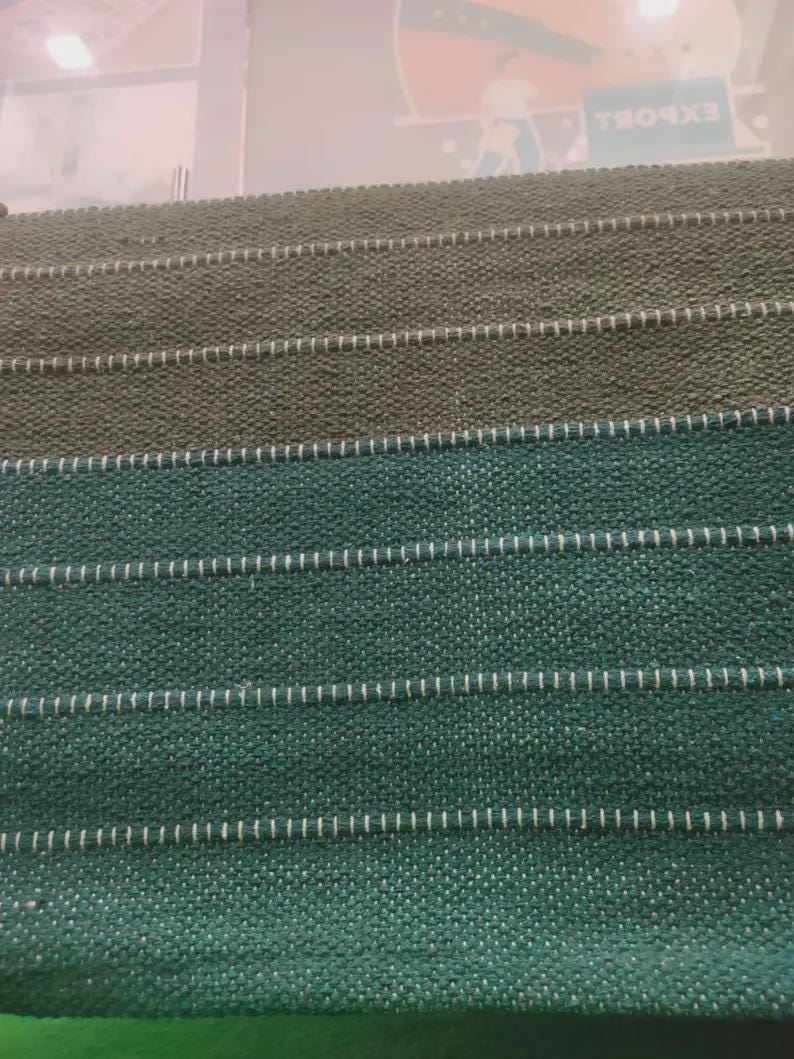 Op de afbeelding: Een stoffenstaalboek met horizontale stroken van getextureerd materiaal in tinten olijfgroen en diep teal. Elke strook wordt gescheiden door een dunne, witte, horizontale stiklijn, wat zorgt voor een gelaagd, rasterachtig uiterlijk.