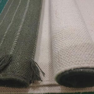 Op de afbeelding: Een donkergroen kleed met witte verticale strepen en franjes aan de randen, naast een opgerold, gebroken wit, getextureerd kleed. Beide kleden hebben een geweven constructie met zichtbare stiksels langs de randen en een dicht, uniform weefsel.