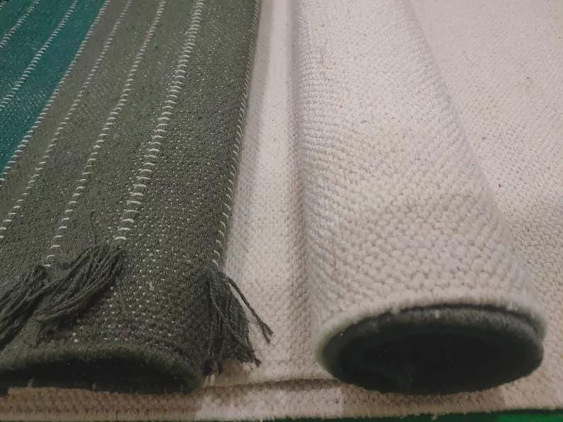 Op de afbeelding: Een donkergroen kleed met witte verticale strepen en franjes aan de randen, naast een opgerold, gebroken wit, getextureerd kleed. Beide kleden hebben een geweven constructie met zichtbare stiksels langs de randen en een dicht, uniform weefsel.