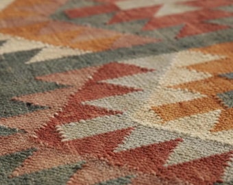 Op de afbeelding: Een geweven vloerkleed met een herhalend geometrisch patroon van driehoeken en ruiten in tinten terracotta, gebrand oranje, crème en leigrijs. Het textiel heeft een getextureerd, geknoopt oppervlak met een consistente poolhoogte.