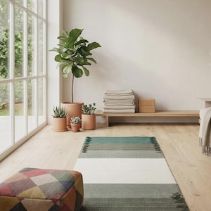Op de afbeelding: Een rechthoekig vloerkleed met horizontale strepen in tinten groen, grijs en een brede witte middenbaan, afgewerkt met korte franjes aan beide uiteinden. Daarnaast staat een vierkante poef met een veelkleurig ruitpatroon.